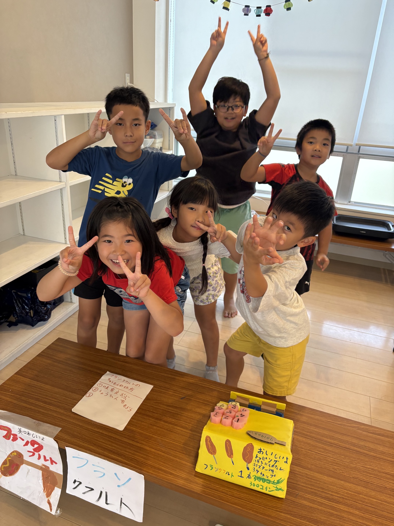 手作りのフランクフルト売り場や看板の後ろでポーズをとる子どもたち